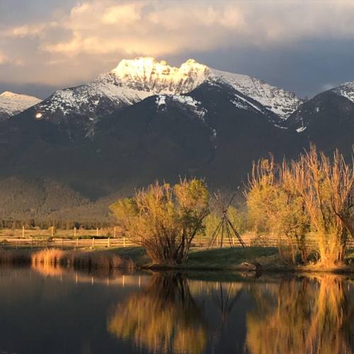 A serene landscape featuring snow-capped mountains, lush greenery, and a calm pond reflecting the scenery at sunset.