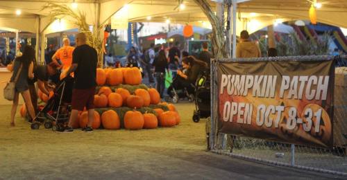 A pumpkin patch with a large stack of pumpkins and a sign announcing it's open from October 8 to 31, with people nearby.