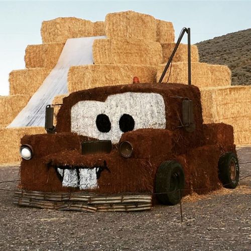 A large sculpture of a truck made from hay bales, featuring a smiling face and set against a backdrop of hay stacks.