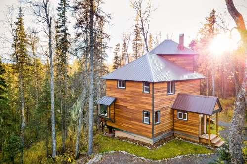 A modern wooden house surrounded by tall trees, with sunlight filtering through the leaves in a serene forest setting.