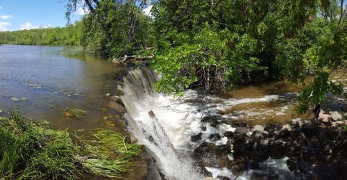 A serene river scene featuring a small waterfall surrounded by lush greenery and rocks under a clear blue sky.