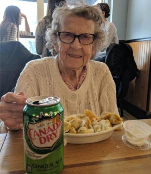 An elderly woman in glasses smiles while holding a can of Canada Dry ginger ale, with a plate of food in front of her.