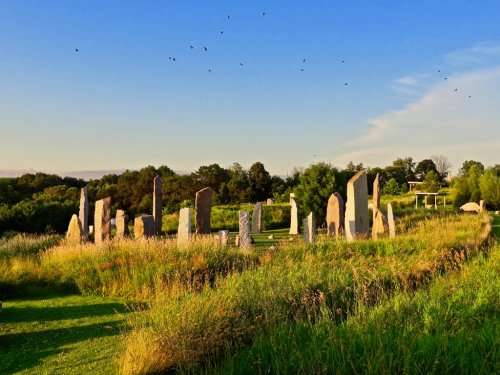 A circle of tall stone monoliths surrounded by lush green grass under a clear blue sky with birds flying above.