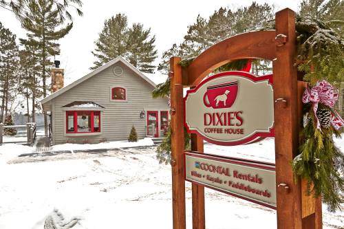 A cozy coffee house sign in winter, surrounded by snow and pine trees, with a festive wreath on the post.