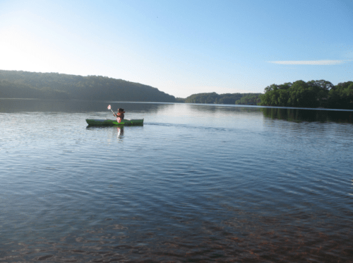 A person paddles a green kayak on a calm lake surrounded by trees and hills under a clear blue sky.