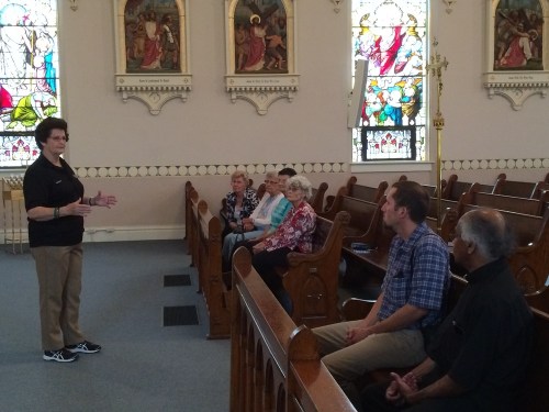 A speaker addresses a small group seated in a church with stained glass windows in the background.
