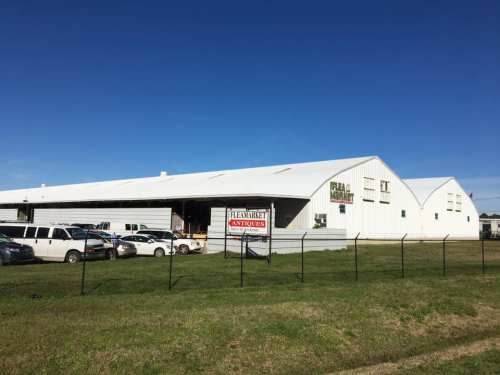 A large white warehouse with a sign reading "Flea Market Antiques," surrounded by parked cars and a clear blue sky.