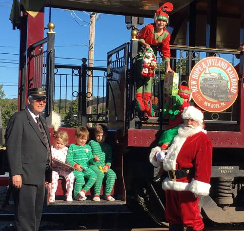 A conductor and Santa pose with children in festive pajamas at a train station decorated for the holidays.