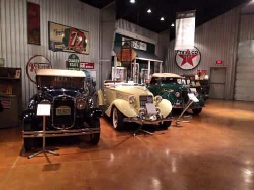Three vintage cars displayed in a museum setting, with retro signage and memorabilia in the background.