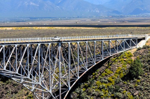 A large metal bridge spans a canyon, surrounded by mountains and open land under a clear blue sky.
