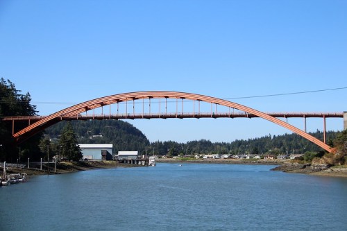 A curved orange bridge spans a calm river, surrounded by trees and buildings under a clear blue sky.