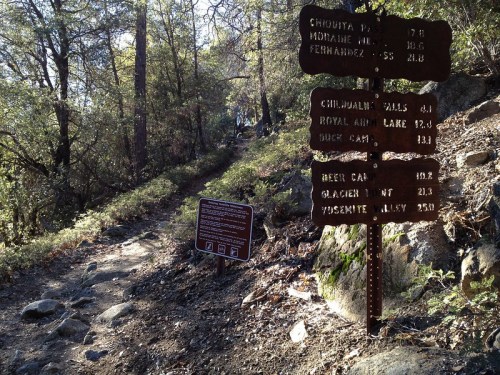 Trail signpost with distance markers for various destinations, surrounded by trees and rocky path.