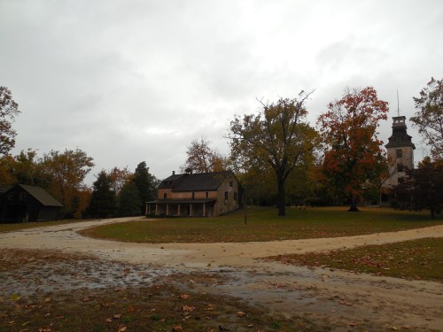 A cloudy day at a rural scene featuring a house, trees with autumn leaves, and a distant clock tower.