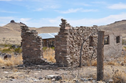 Ruins of a stone building in a dry landscape, with distant hills and a clear blue sky.