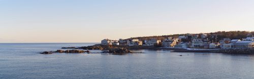 Coastal view of a serene shoreline with houses, rocky outcrops, and calm waters under a clear sky.