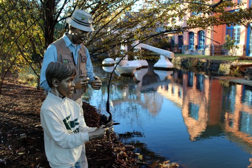 A statue of a man teaching a boy to fish by a serene river, surrounded by trees and colorful buildings in the background.