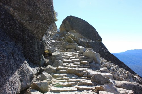 A rocky path leads up a steep slope, flanked by large boulders and offering a view of distant mountains.