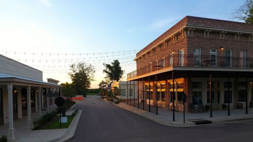 A charming street scene at sunset, featuring brick buildings and string lights, with trees lining the road.