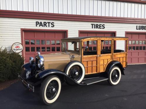 A vintage car with a wooden body parked in front of a garage with red doors and a sign for parts and tires.