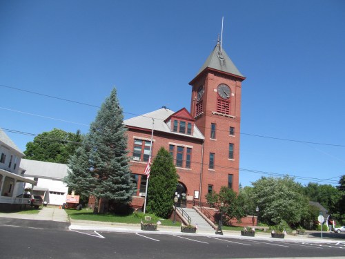 Historic red brick building with a clock tower, surrounded by trees and a clear blue sky. American flag visible.