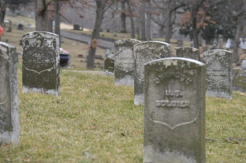 Gravestones in a cemetery, some engraved with "U.S. Soldier," surrounded by grass and trees.