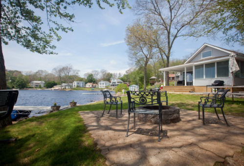 A lakeside scene featuring a fire pit surrounded by chairs, with a house and trees in the background.