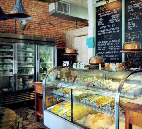 A cozy bakery interior featuring a glass display case filled with various desserts and a chalkboard menu on the wall.