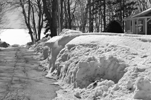 A snowy road lined with large snowbanks, leading towards a frozen lake in a black and white landscape.