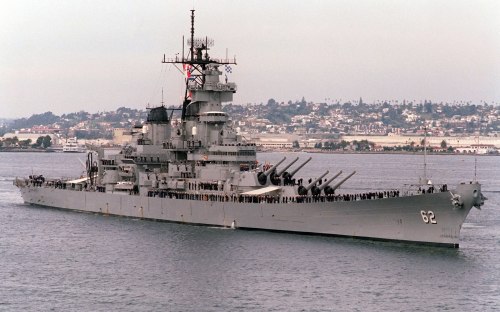A large battleship with multiple gun turrets sails in calm waters, with a city skyline in the background.