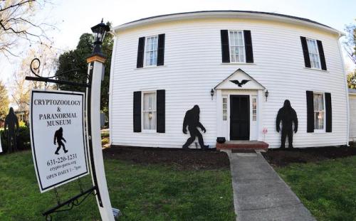 A white house with black shutters and silhouettes of Bigfoot outside, featuring a sign for a cryptozoology museum.
