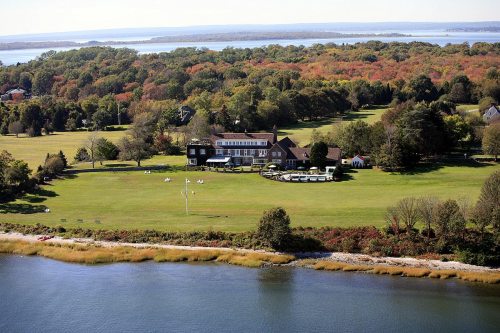 Aerial view of a large estate with a green lawn, surrounded by trees and a body of water, under a clear blue sky.