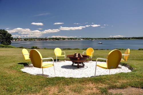 A fire pit surrounded by yellow chairs on a grassy area, overlooking a calm lake and blue sky with clouds.