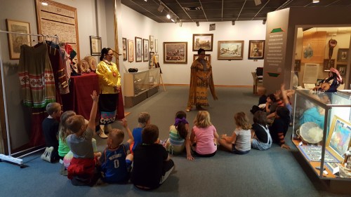 Children sitting on the floor in a museum, watching two performers in traditional attire. Artifacts and displays are visible.