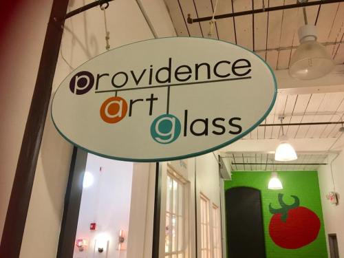 Sign for "Providence Art Glass" hanging in a brightly lit space with a green wall featuring a tomato design.