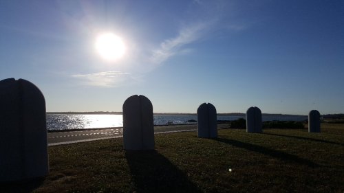 A sunny landscape featuring large, rounded sculptures on a grassy area by the water, with a clear blue sky.