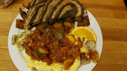 A plate of scrambled eggs topped with vegetables and sauce, served with roasted potatoes, orange slice, and toasted bread.