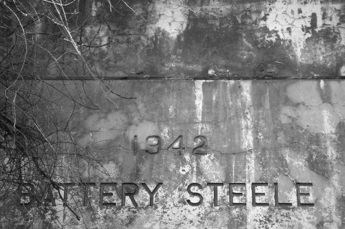 Weathered wall with the text "1942 BATTERY STEELE" and bare branches above, in black and white.