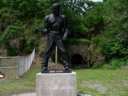 A black statue of John Henry holding a hammer, set against a green hillside and a tunnel entrance in the background.