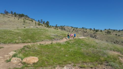 A group of five hikers walking along a grassy trail in a sunny, mountainous landscape.
