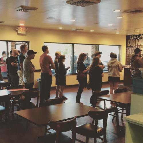 A line of people waiting inside a cafe, with tables and large windows in the background.