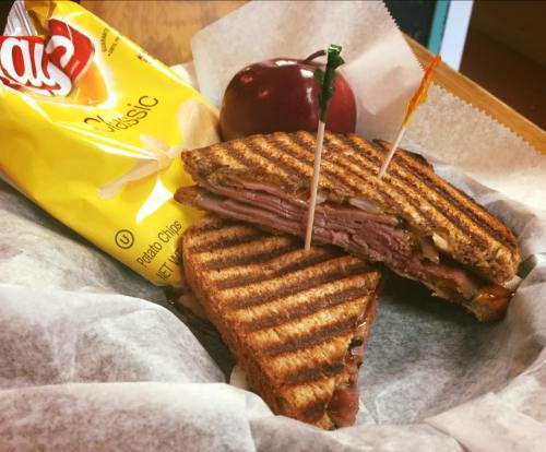 A grilled sandwich with layers of meat, served with potato chips and a red apple, all in a paper-lined basket.