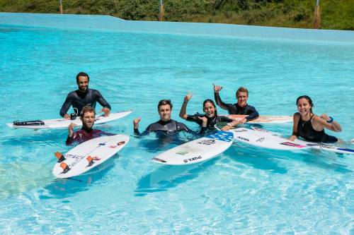 A group of six surfers in a pool, smiling and posing with surfboards, enjoying a sunny day.
