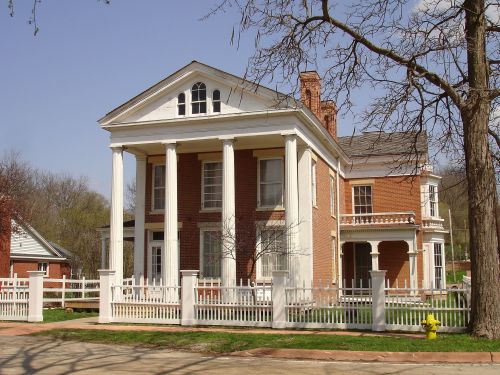 A historic brick house with white columns, a porch, and a picket fence, surrounded by trees and a clear blue sky.