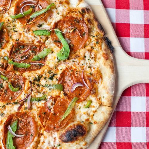 A close-up of a pepperoni pizza topped with green peppers and red onions, on a wooden pizza peel over a checkered tablecloth.