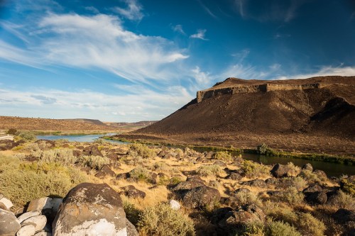 A scenic landscape featuring a river, rocky terrain, and a distant hill under a blue sky with wispy clouds.
