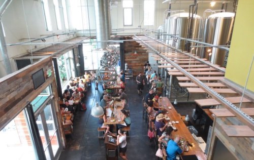 A bustling brewery interior with patrons at the bar and tables, large brewing tanks in the background, and a modern design.