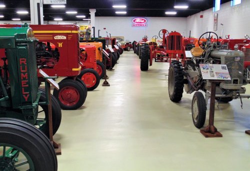 A row of vintage tractors in a museum, showcasing various models and colors under bright lighting.
