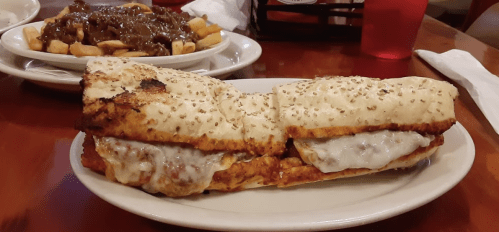 A large sandwich with melted cheese on toasted sesame seed bread, served alongside a plate of poutine.