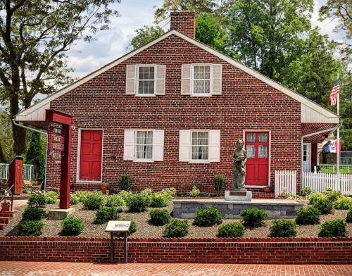 A brick house with red doors and shutters, surrounded by greenery and a statue in the front yard.