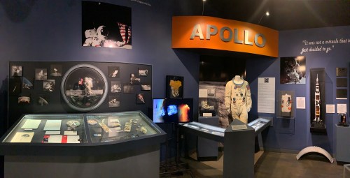 Exhibit showcasing Apollo mission artifacts, including photos, a spacesuit, and a model rocket in a museum setting.